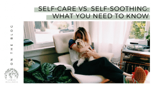 Self-Care vs. Self-Soothing: What You Need To Know (A woman sits on a white chair in a brightly light room featuring plants and midcentury decor. she is hugging a small white dog on her lap.)