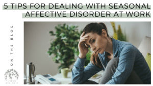 A graphic that reads "5 Tips for Dealing With Seasonal Affective Disorder at Work" Above a stock photo of a white woman sitting at a desk with her head in her hands, looking distressed.